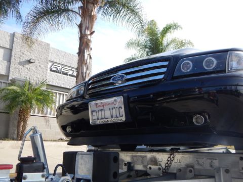 2003 Ford Crown Victoria - front of a black Ford sedan shows halo headlights and a California EVIL VIC plate with a Crown Victoria frame while sitting on a trailer.