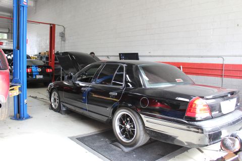 2003 Ford Crown Victoria - black sedan with its hood open and black aftermarket wheels sits inside an auto shop beside a blue two-post lift and red-striped wall.