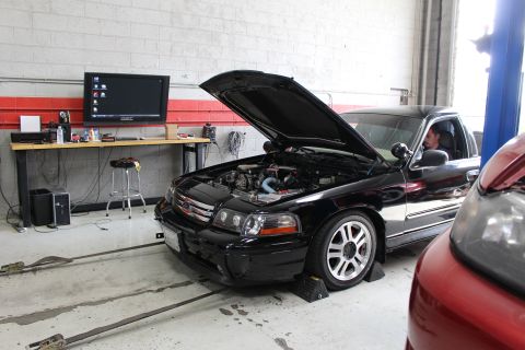 2003 Ford Crown Victoria - black Ford sedan with its hood up sits strapped on a dyno inside a shop, a tuning monitor glowing on the workbench behind it.
