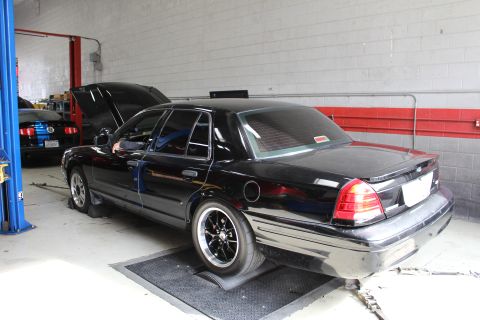 2003 Ford Crown Victoria - Rear three-quarter view of a black sedan on black wheels with its hood raised, parked on a dyno mat inside a gray-walled auto shop.