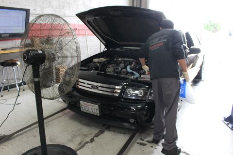 2003 Ford Crown Victoria - mechanic in a green jacket leans over the engine bay of a black Ford on a dyno, a large floor fan blowing toward the EVIL VIC front.