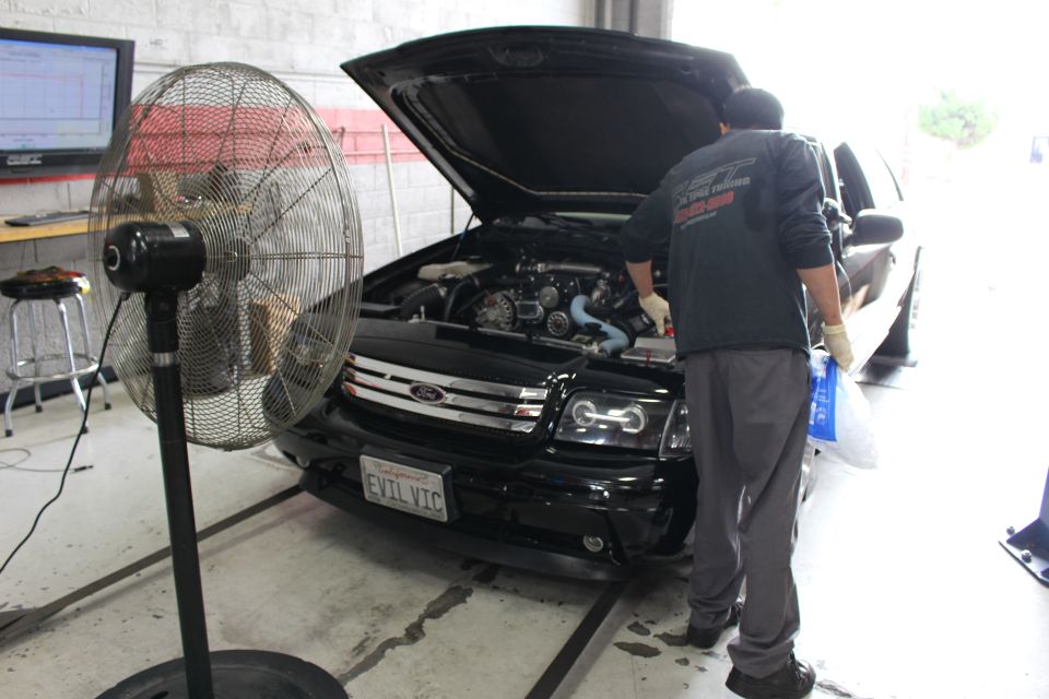 Mechanic in green jacket working under the hood of a black Ford on a dyno with a fan