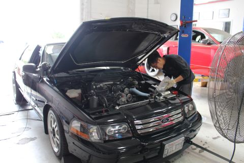 2003 Ford Crown Victoria - mechanic reaches into the engine bay of a black Ford sedan with halo lights and an EVIL VIC plate while a fan stands beside the car.