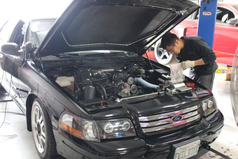 2003 Ford Crown Victoria - mechanic works inside the engine bay of a black Ford sedan with halo headlights and an EVIL VIC California plate in an auto shop.