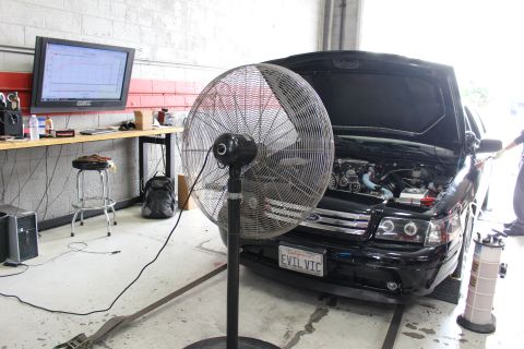 2003 Ford Crown Victoria - black Ford sedan with EVIL VIC plate and open hood sits on a dyno behind a big floor fan, a tuning monitor glowing on the bench.