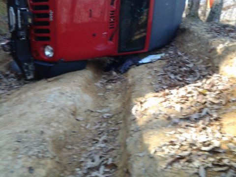 1995 Ford Bronco - An overturned red SUV lies on its side on a dirt trail, surrounded by fallen leaves and trees.