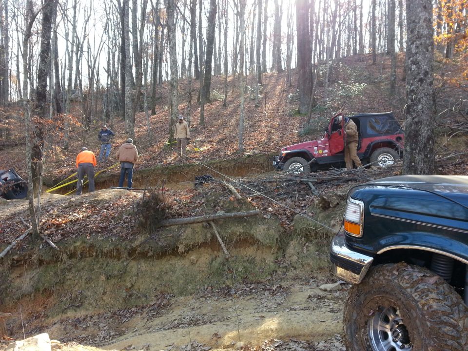 Dark green truck in foreground, red Jeep partially stuck on embankment, people working with yellow straps in wooded area