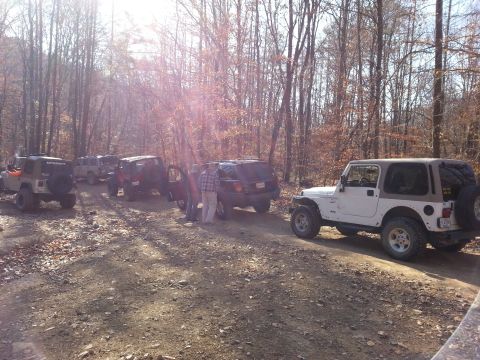 1995 Ford Bronco - A group of off-road vehicles are parked on a dirt road surrounded by trees in autumn.