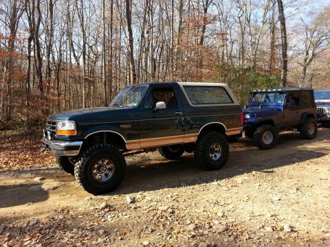 1995 Ford Bronco - A lifted 1995 Ford Bronco with tan trim is parked on a dirt road, followed by other off-road vehicles.