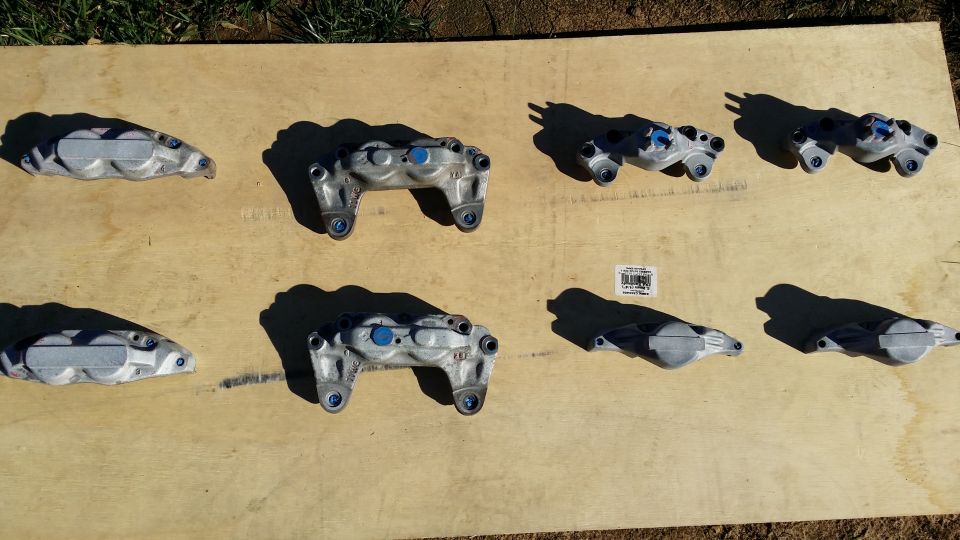 Eight brake calipers laid out on a wooden surface. Some have blue accents
