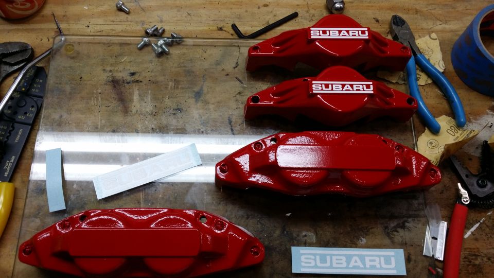 four red Subaru brake calipers with "SUBARU" decals, tools, and "SUPERMOTORS" sticker on a workbench