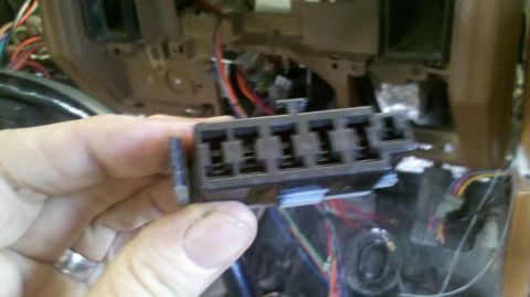 1982 Ford Bronco - A hand holds a black plastic automotive fuse block with six slots. The fuse block is positioned in front of a partially disassembled dashbo…