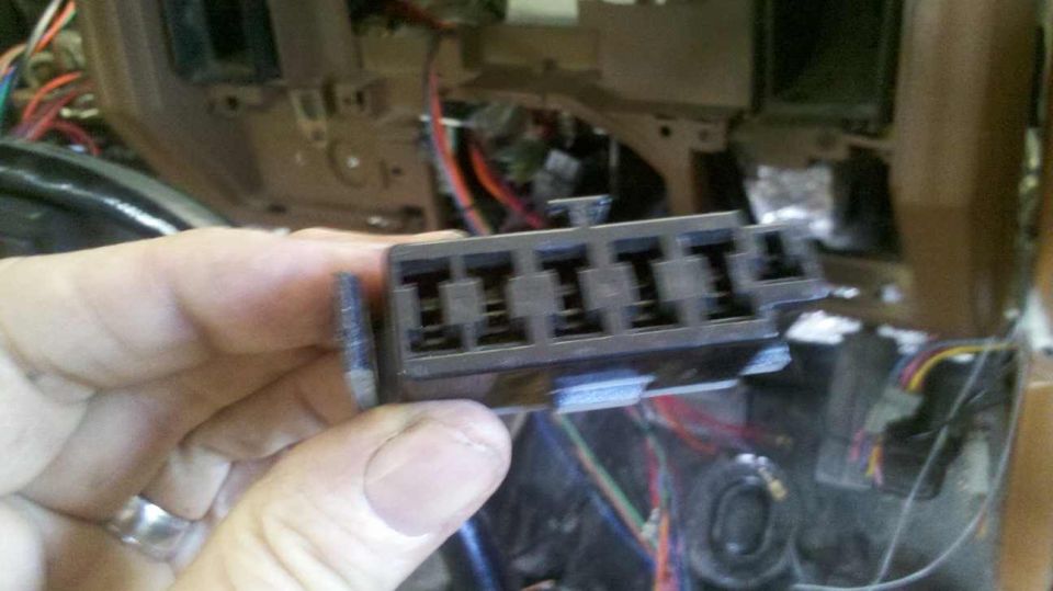 Close-up of a hand holding a black fuse block with six slots, in front of car dashboard wiring.