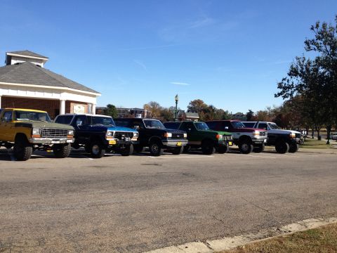 1974 Ford Bronco - A line of lifted pickup trucks and SUVs are parked in a lot in front of a building. The vehicles are various colors including yellow, blue,…