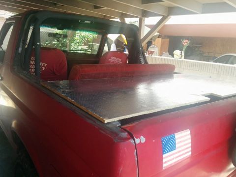 1988 Ford Bronco - The rear passenger side of a red Ford Bronco is shown, covered with a makeshift wooden bed cover. An American flag sticker is visible on th…
