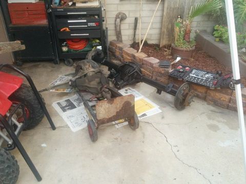 1988 Ford Bronco - Two vehicle rear axle assemblies are laid out on the ground next to a red ATV. A toolbox and various tools are visible on a brick wall in t…