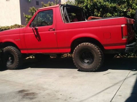 1988 Ford Bronco - The side profile of a red Ford Bronco is shown parked on a concrete driveway. The vehicle has a soft top removed, exposing the interior and…