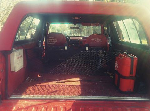 1988 Ford Bronco - The interior of a red Ford Bronco is shown, looking towards the front seats and dashboard. The cargo area contains a first aid kit, a black…