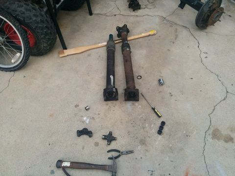 1988 Ford Bronco - Two driveshafts and associated parts, including a U-joint and a hammer, are laid out on a concrete surface next to a tire and wheel assembl…