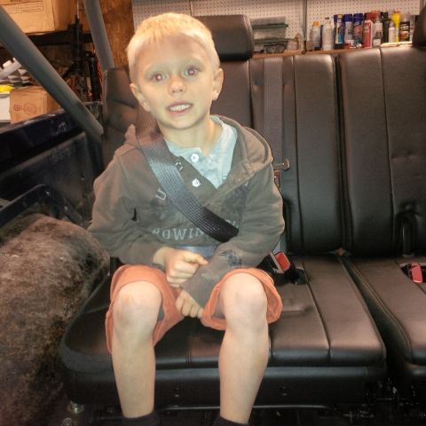 1978 Ford Bronco - A young boy with blonde hair and red-tinted eyes sits in the back seat of a vehicle, wearing a grey shirt and a brown hoodie. He is wearing…