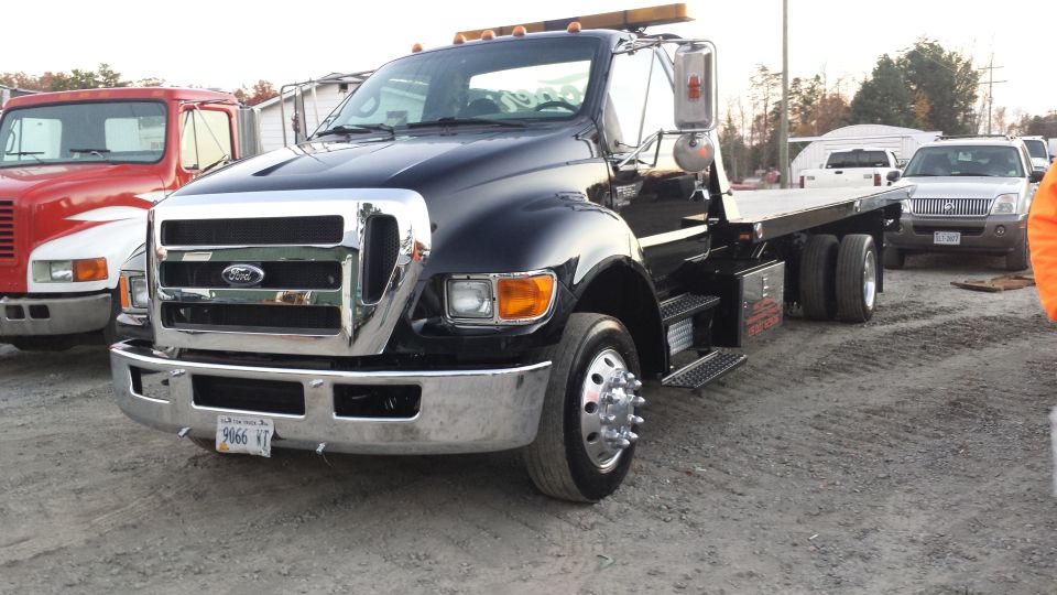 Black Ford F-650 tow truck with chrome grille and flatbed, parked on gravel.