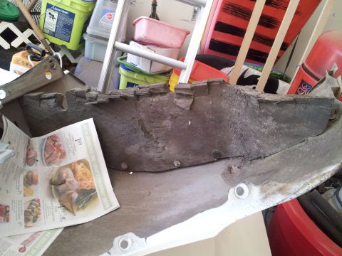 1976 Ford Ranchero - A damaged, weathered car bumper part is shown resting on a newspaper. The background is cluttered with various household items and storage…