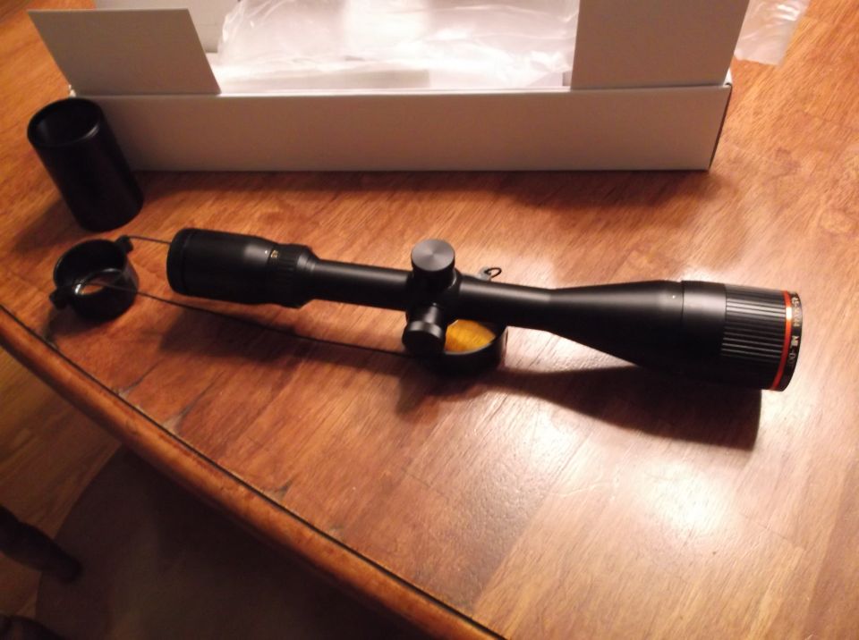 Black rifle scope with lens cap and mount on a wooden table.