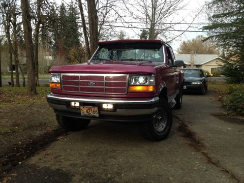 1992 Ford F-150 - The front view of a dark red Ford F-150 pickup truck is parked on a paved driveway. The truck has a chrome grille, clear headlights, and a…