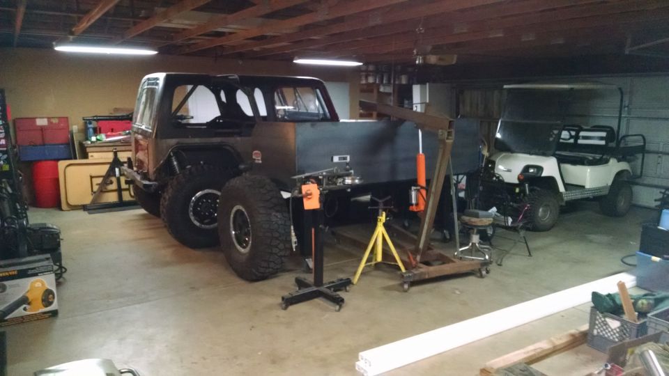 Modified Jeep CJ7 with large tires and a custom bed in a garage, next to a white golf cart.