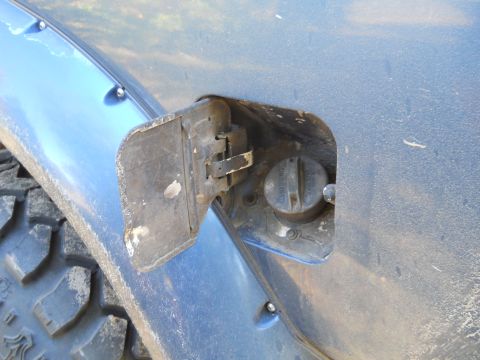 1992 Ford Bronco - Close-up view of an open fuel filler door on a blue vehicle, revealing the gas cap and surrounding area. The vehicle has a tire and fender…