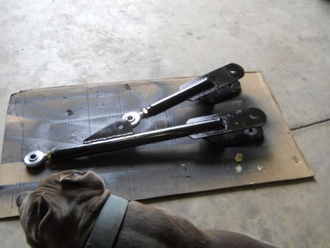 1992 Ford Bronco - Two black painted suspension control arms with adjustable ends are laid out on a piece of cardboard on a concrete floor. A dog's head is vi…