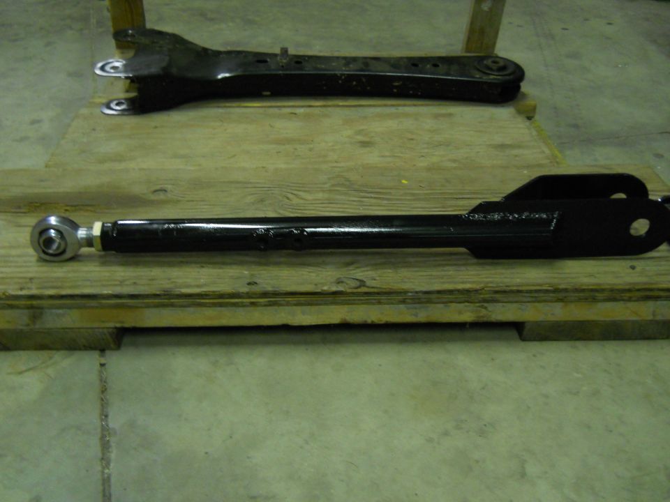 Two black suspension control arms on a wooden pallet. One is in the foreground with a spherical joint.