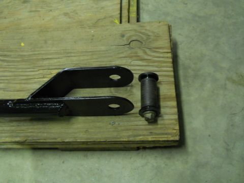 1992 Ford Bronco - A black metal bracket with two oval holes is shown alongside a bolt with a dark gray cylindrical bushing. Both items are resting on a woode…