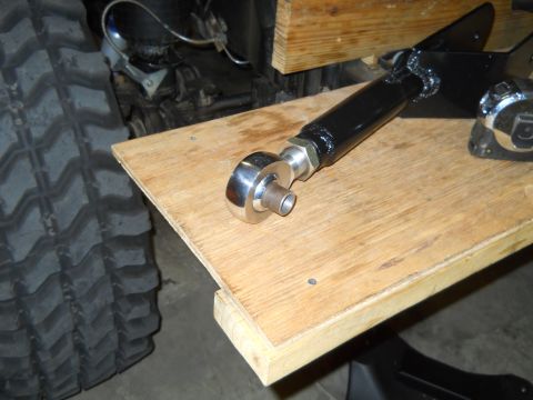 1992 Ford Bronco - A close-up view of a black adjustable control arm with a silver heim joint resting on a wooden workbench. A large, knobby tire is visible t…