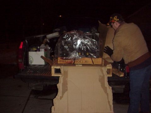 1989 Ford Bronco - A person is loading a large engine, wrapped in plastic, from a wooden pallet into the bed of a pickup truck at night.