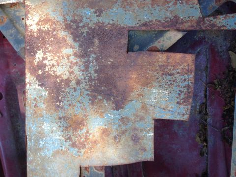 1979 Ford Bronco - This image shows a close-up of a heavily rusted metal panel, likely from a vehicle's body, with peeling blue paint revealing red primer und…
