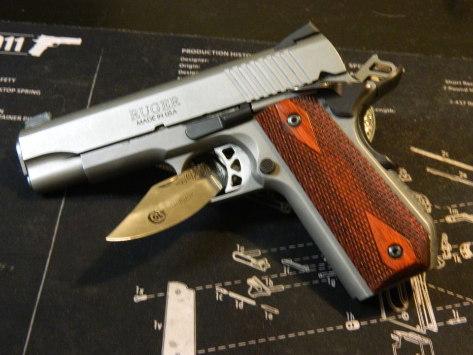 Silver Ruger pistol with wood grips and a partially opened knife inserted into the trigger guard, resting on a black mat with diagrams