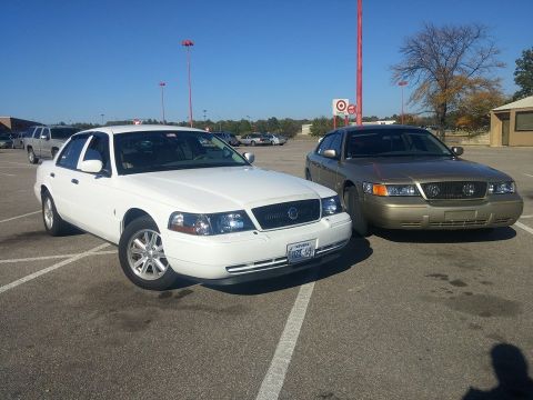 2000 Mercury Grand Marquis - Two Mercury Grand Marquis sedans are parked side-by-side in a parking lot under a clear blue sky.