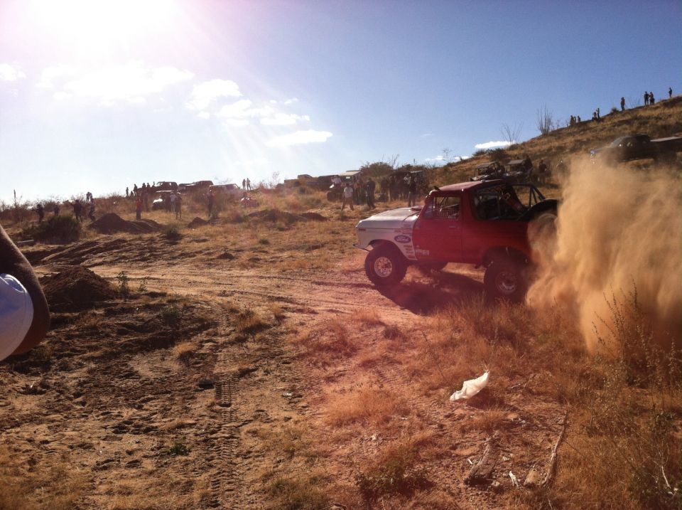 Red and white Ford Bronco kicking up dust on an off-road course with spectators on a hill in the background