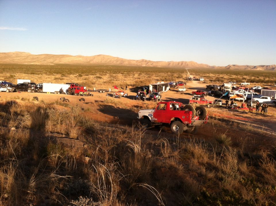 Red off-road truck with spare tire on back, driving on dirt path. Many other vehicles and trailers in background desert scene