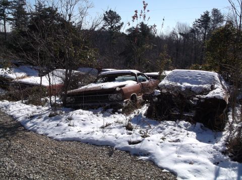 1974 Dodge Challenger - Two old cars are partially buried in snow and brush in a wooded area.