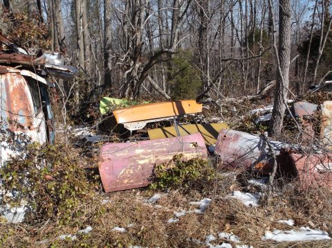 1974 Dodge Challenger - Scattered car parts, including doors and hoods, lie in a wooded junkyard with patches of snow on the ground.
