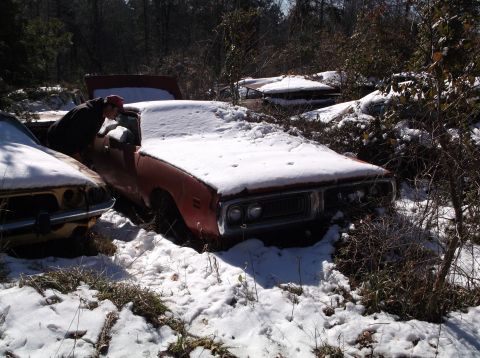 1974 Dodge Challenger - A person inspects a snow-covered 1973 Dodge Charger among other abandoned vehicles in a wooded area.