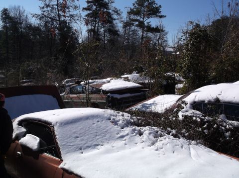 1974 Dodge Challenger - Multiple vehicles sit abandoned in a snowy junkyard, overgrown with vegetation.