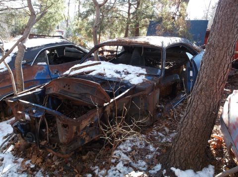 1974 Dodge Challenger - A heavily rusted car sits in a junkyard, partially covered in snow, with trees growing around it.