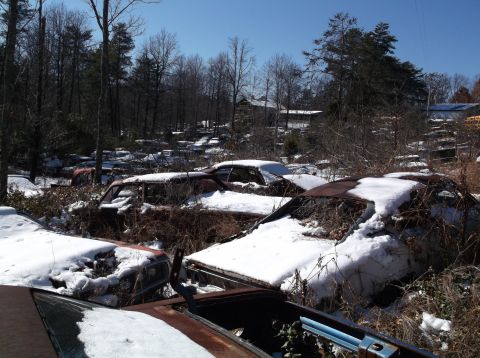 1974 Dodge Challenger - Several old cars are buried in snow at a junkyard, with trees and buildings in the background.