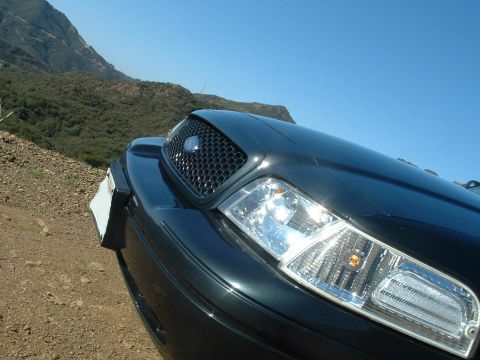 1998 Ford Crown Victoria - Low close-up of a dark sedan's honeycomb-mesh grille and illuminated headlight, set against blue sky and dry hills.