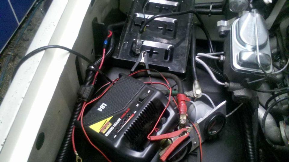 Battery charger with red and black clamps connected to a car battery terminal. Brake fluid reservoir visible