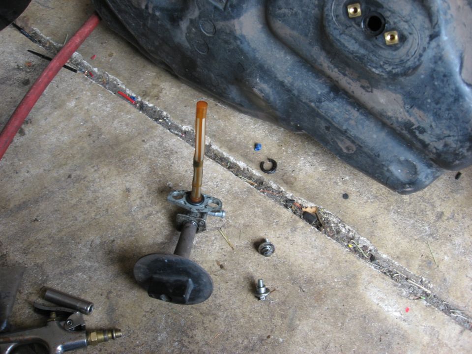 Removed ATV fuel petcock with standpipe filter on a garage floor beside the upturned fuel tank