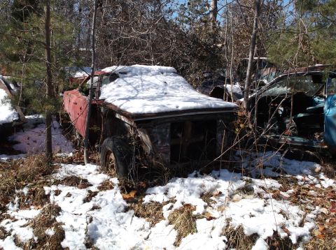 1974 Dodge Challenger - A rusty red vintage car, partially covered in snow, sits in a wooded area. The car's hood and roof are covered in snow, and its front end i…